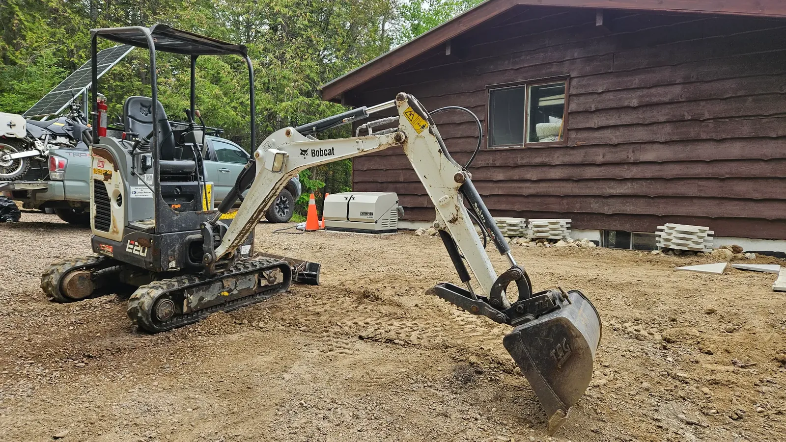 Compact excavator parked near a rural cabin in a wooded setting.