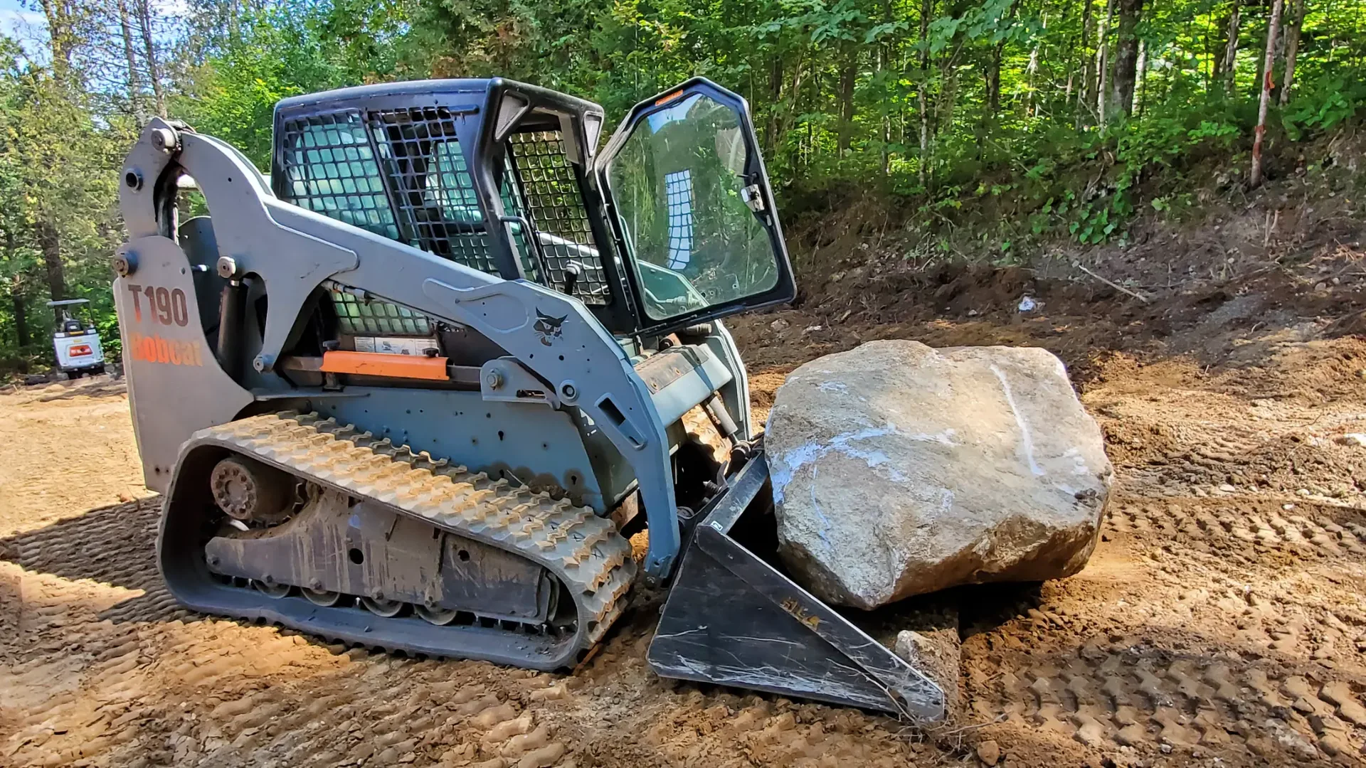 Compact track loader moving a large boulder at a rural site.