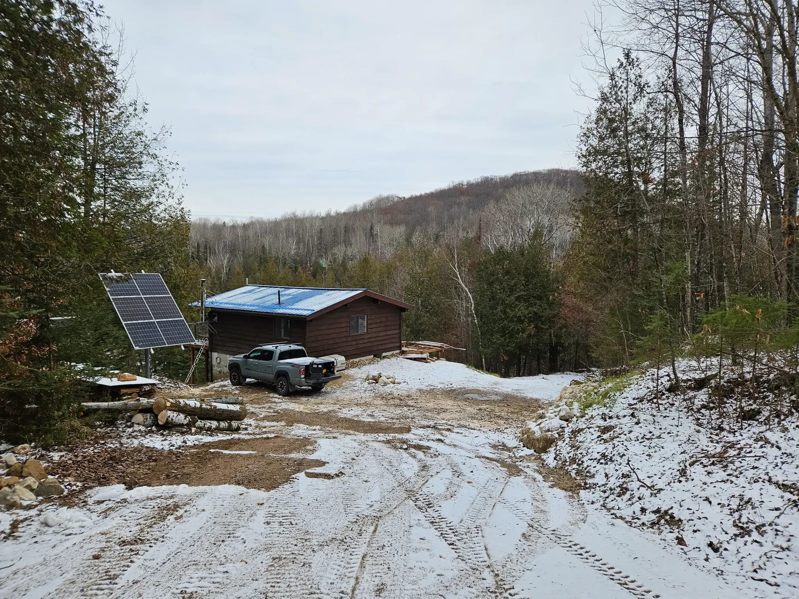 Pole-mounted solar array, vehicle, and cabin in light snow at a wooded rural property.